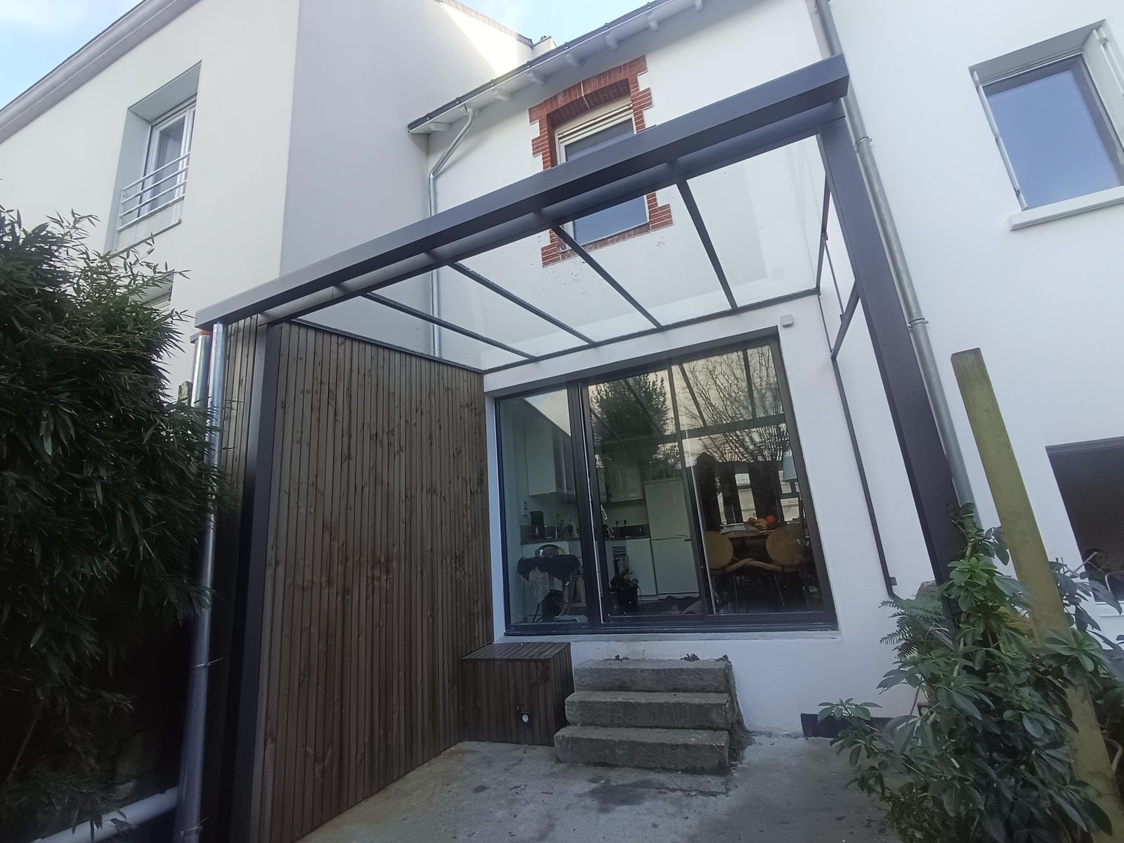 Véranda en acier et verre installée dans une cour, contre une façade blanche.